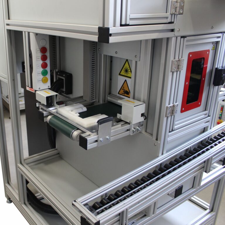 Automatyczna przelotowa znakowarka laserowa w zabudowie przemysłowej, wyposażona w transporter i moduły wizyjne.